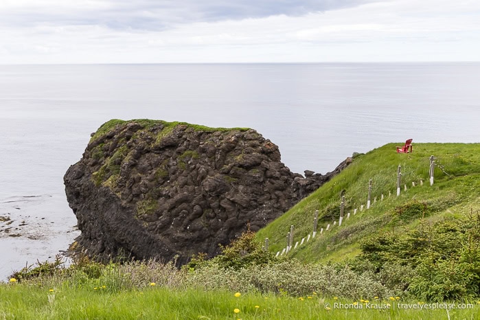 Read chair on a grassy clifftop looking out at a large rock and the Gulf of St. Lawrence. 