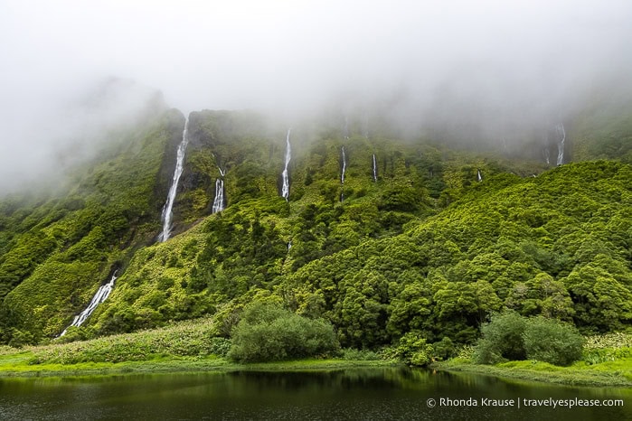 Waterfalls flowing down a lush cliffside into a lake on a foggy day. 